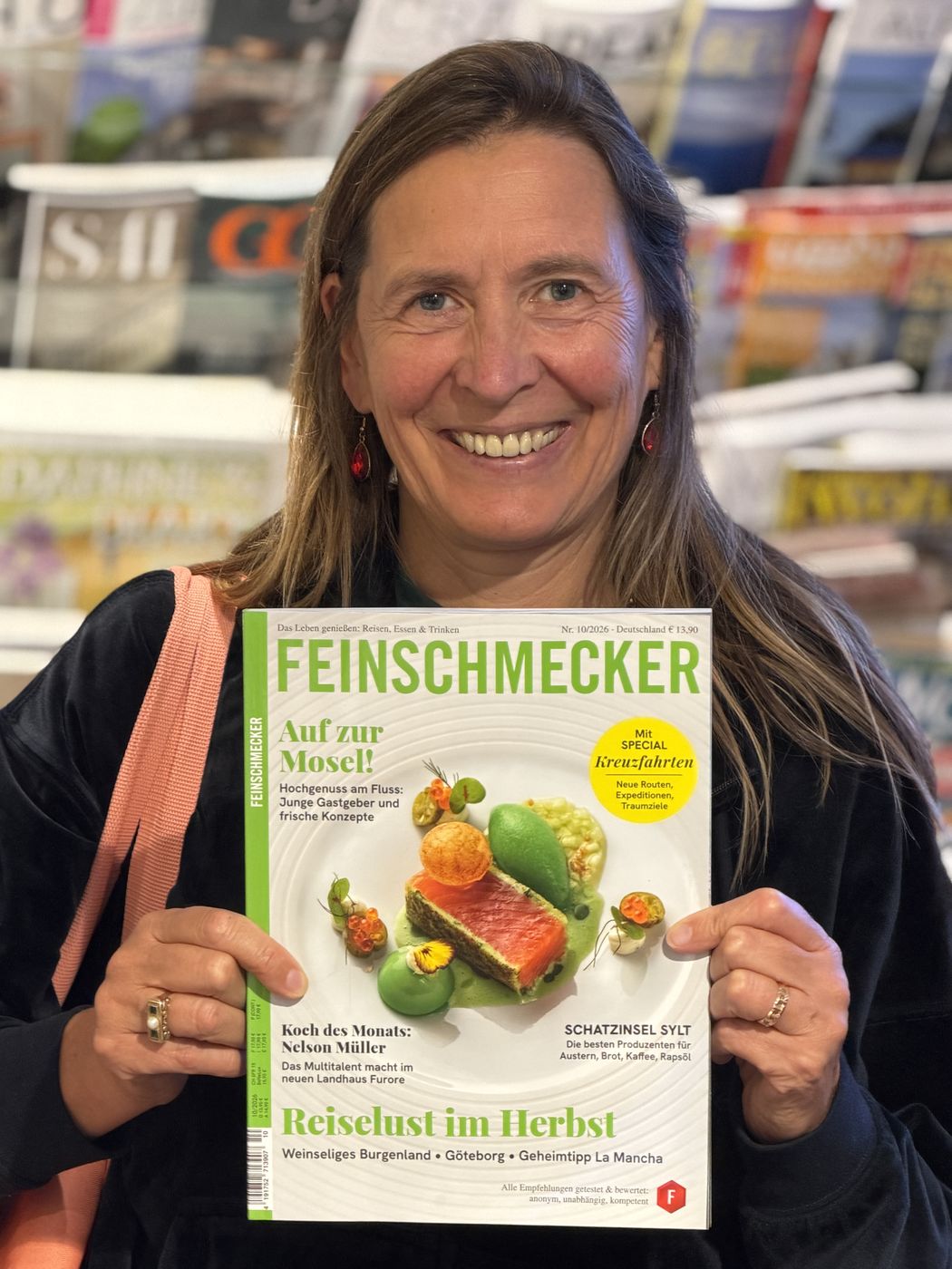 Sabine Ryan hält lächelnd die Zeitschrift Feinschmecker vor einem Zeitschriftenregal